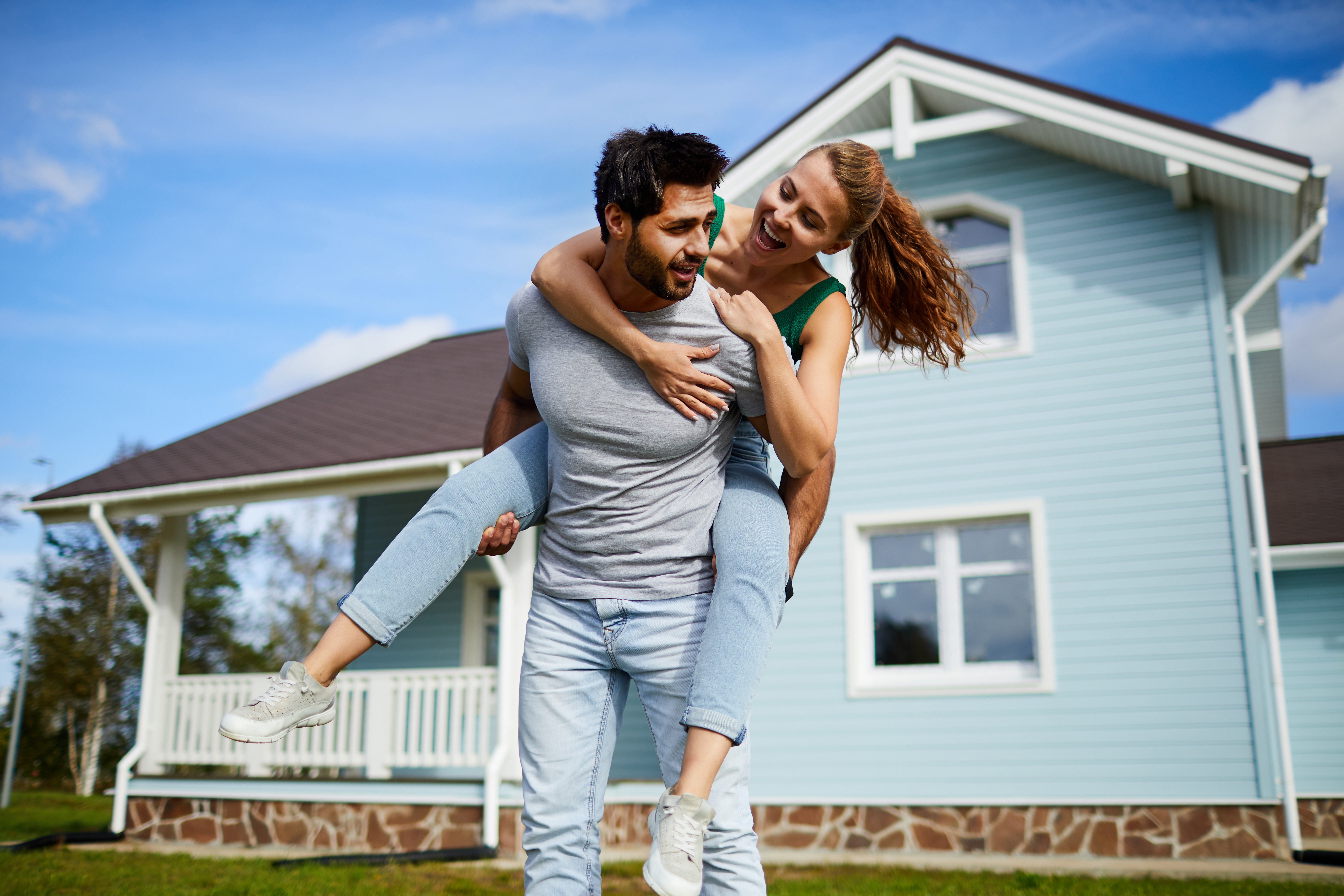 Young man giving his cheerful girlfriend piggyback on background of their new house